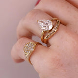 A close-up of a hand wearing two gold rings. The larger ring features a pear-cut clear gemstone, and the smaller ring has a round pavé-set clear gemstone, both on a soft pink background.