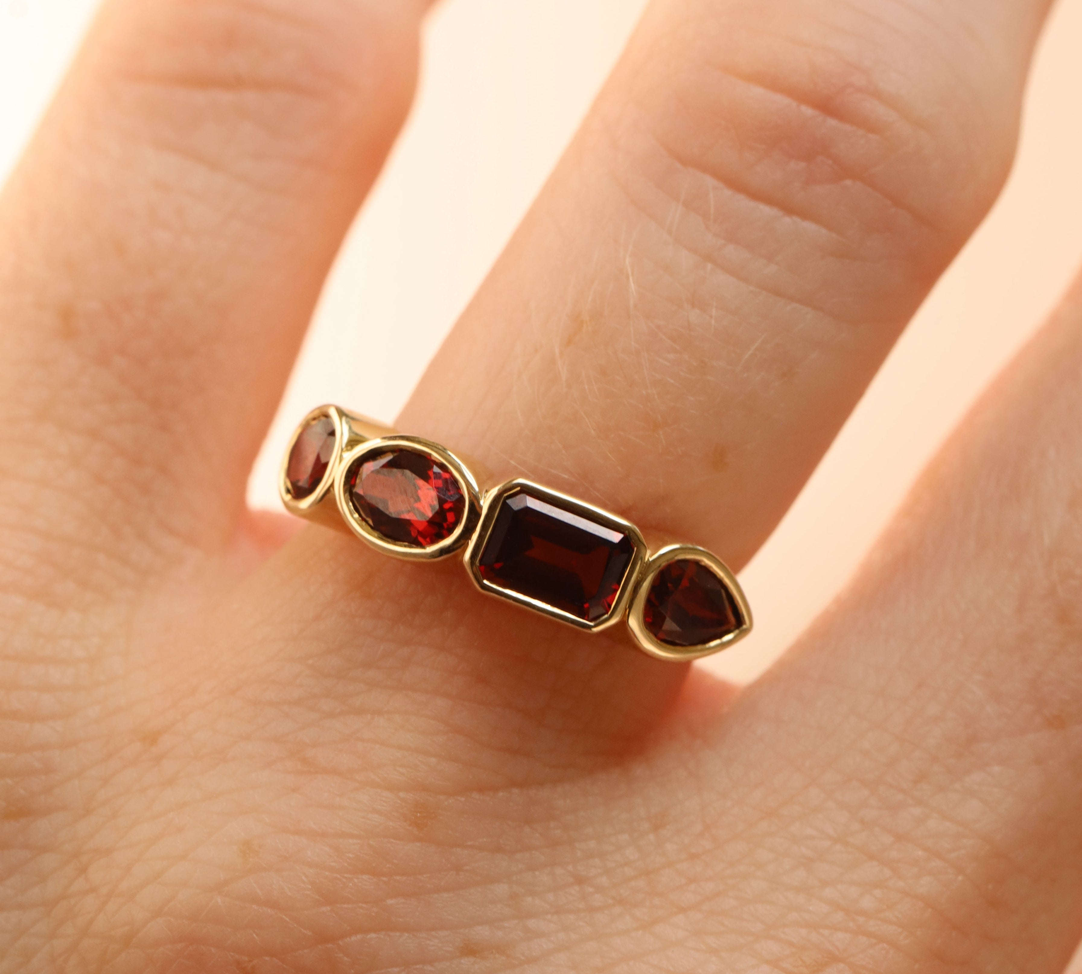 mixed shape garnet bezel band on woman's finger, against a neutral background (a mix of round, emerald step-cut, pear, and oval faceted stones) all set in 14k yellow gold