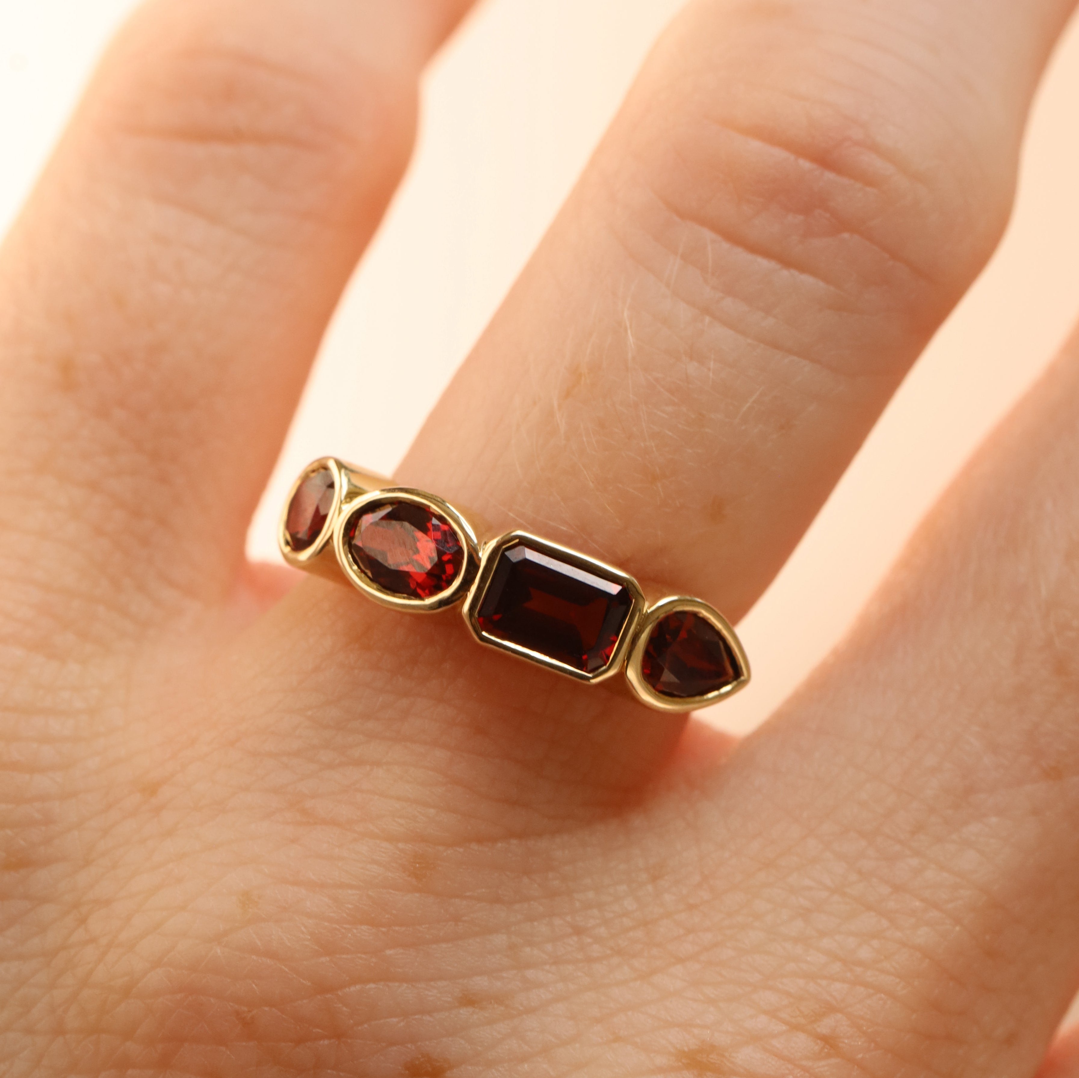 mixed shape garnet bezel band on woman's finger, against a neutral background (a mix of round, emerald step-cut, pear, and oval faceted stones) all set in 14k yellow gold