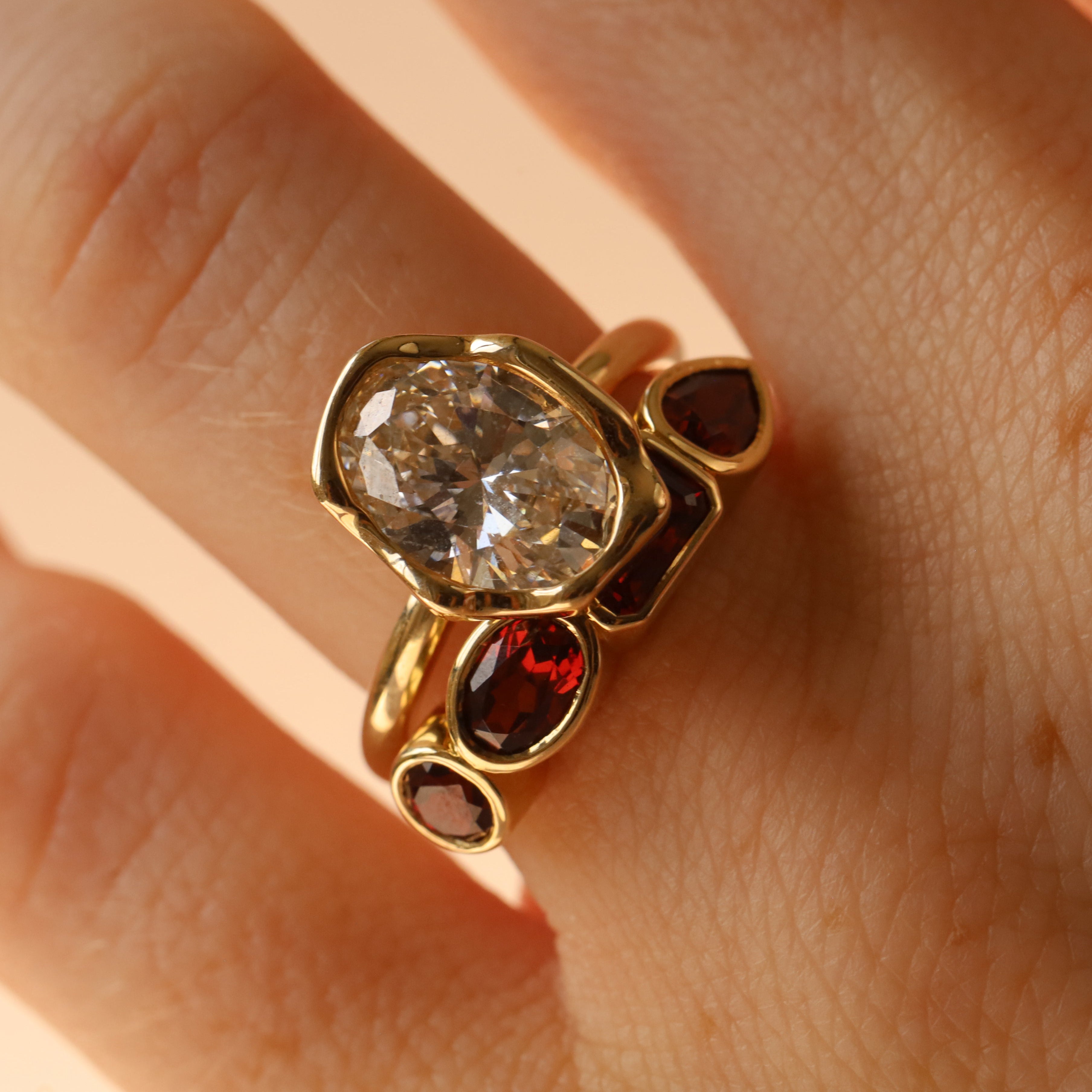 mixed shape garnet bezel band on woman's finger stacked with an oval diamond engagement ring with an abstract bezel, against a neutral background