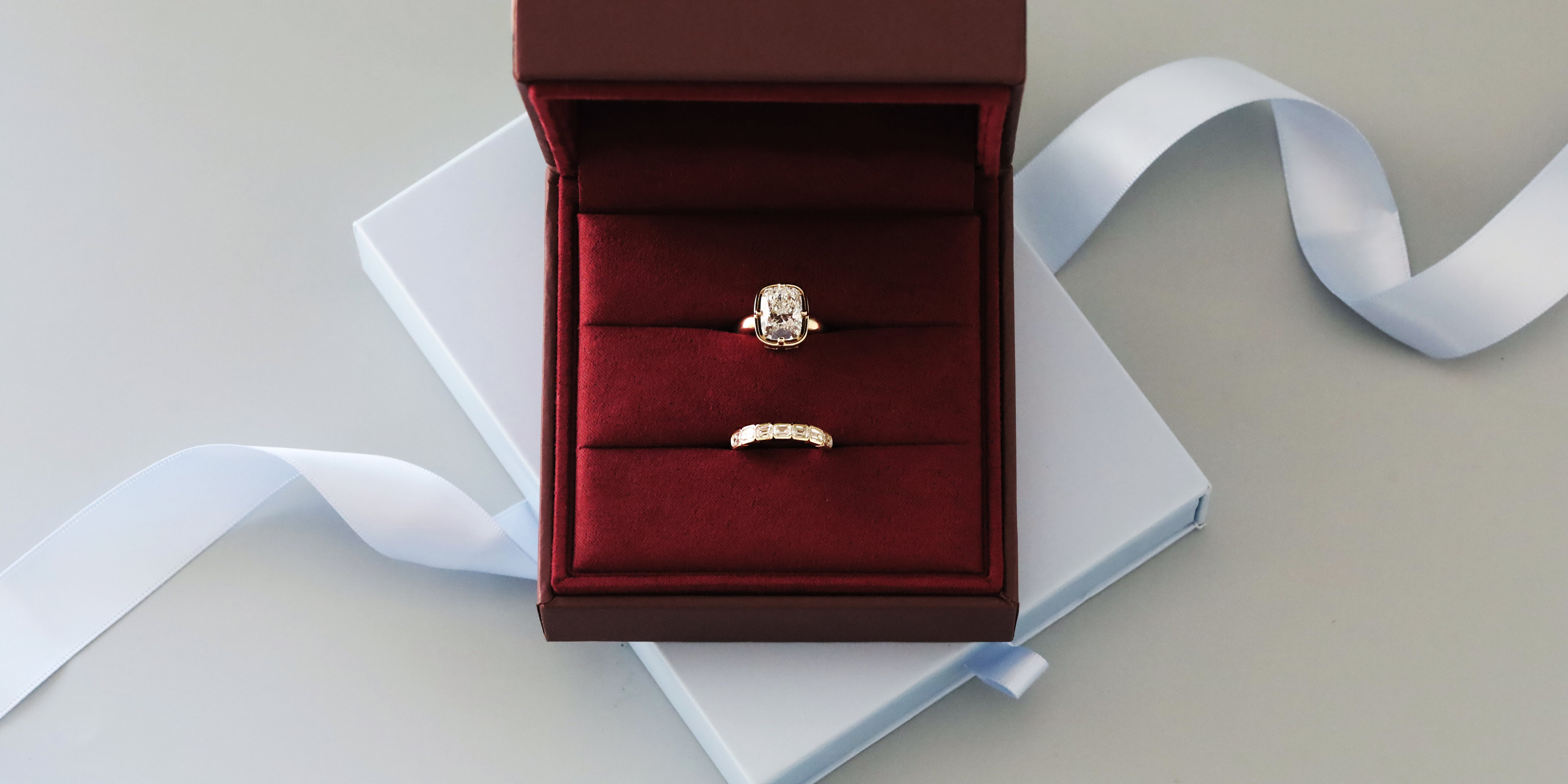 Cushion-cut diamond engagement ring and emerald-cut diamond band in a maroon ring box on a pale blue gift box with satin ribbon