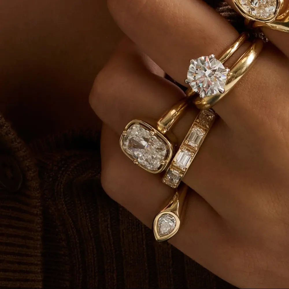A close-up lifestyle shot of a woman wearing multiple gold rings with clear gemstones, a matching gold earring, and a brown ribbed knit top.