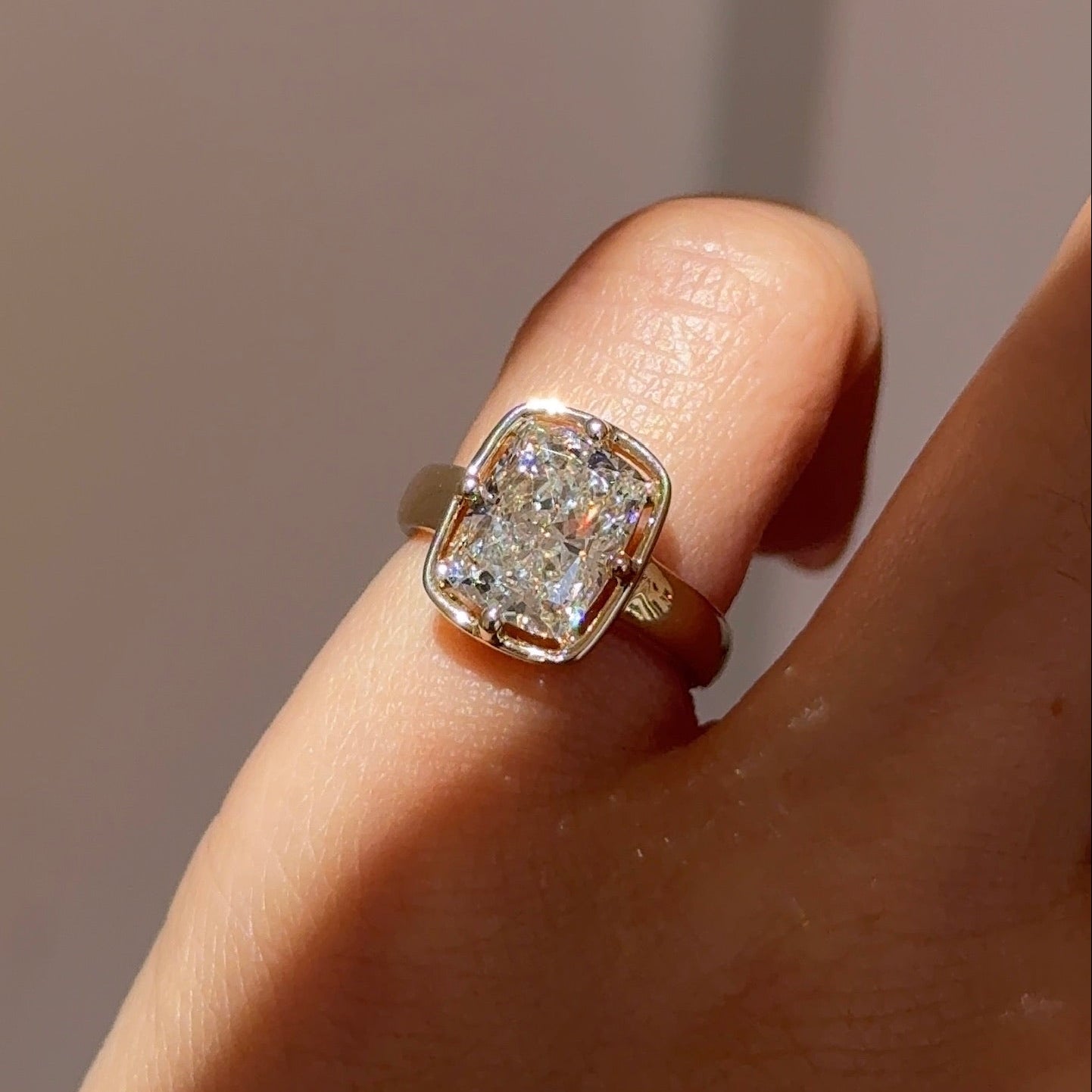 A close-up of a hand wearing a yellow gold ring with a cushion-cut clear gemstone, set against a soft, warm-toned neutral background.