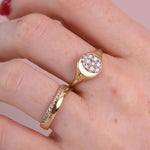 A close-up of a hand wearing a gold ring with a round pavé-set clear stone and a slim matching gold band with small clear stones, against a soft pink background.
