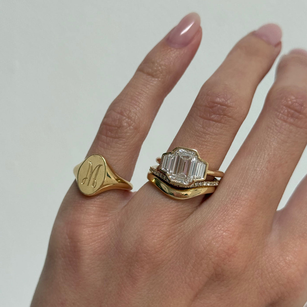 A close-up of a hand wearing two gold rings. The ring on the middle finger features a large emerald-cut clear center stone with smaller clear stones, while the ring on the ring finger is a polished yellow gold signet with an engraved script letter 'M'.