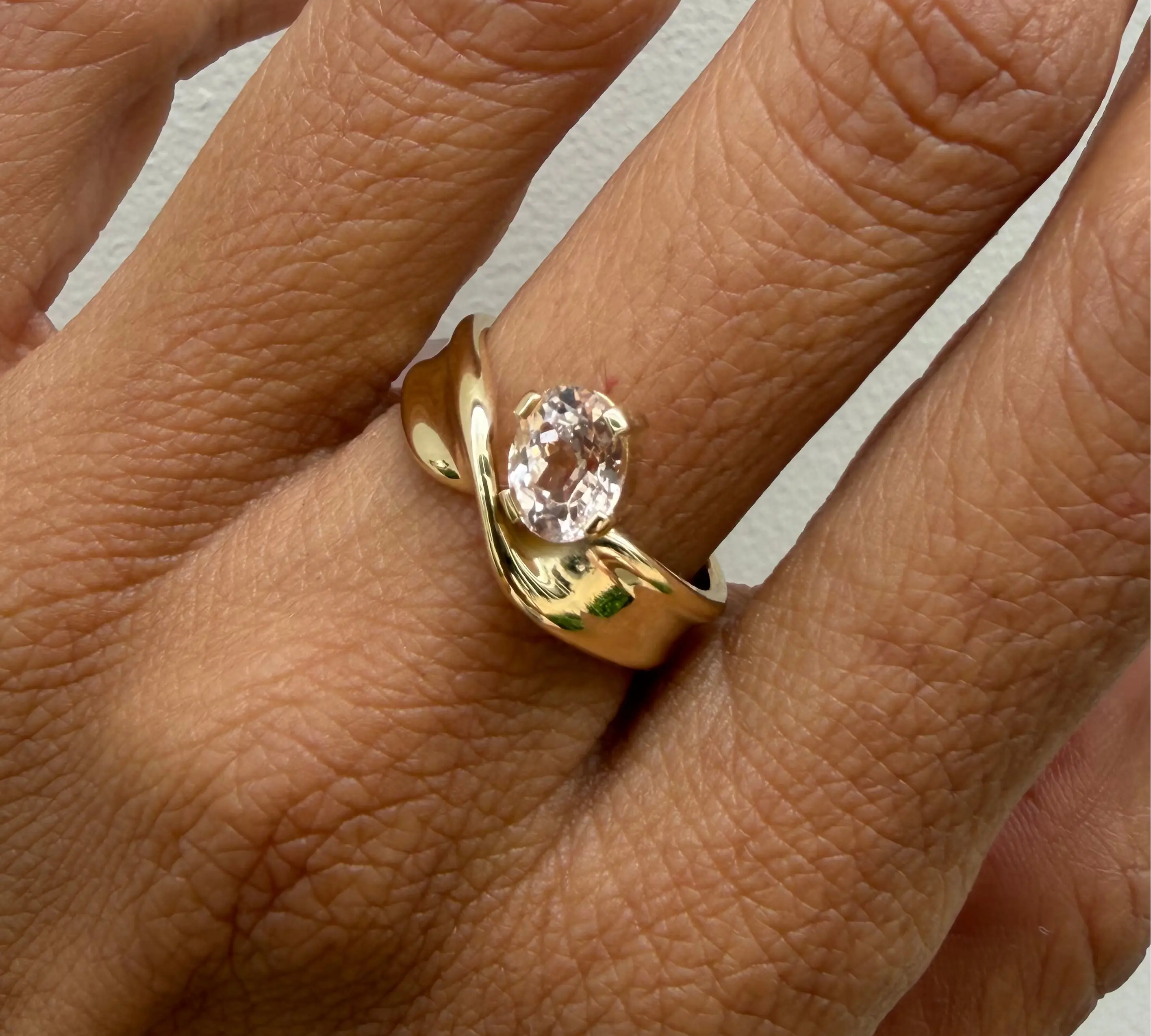 Gold ring with a peach sapphire on a person's finger against a neutral background