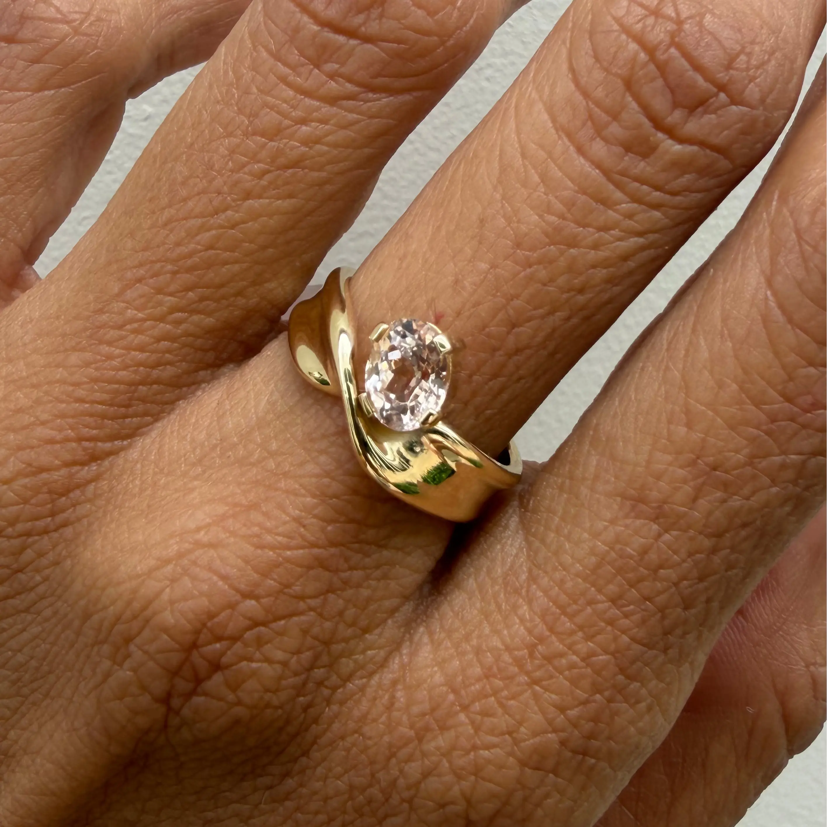Gold ring with a peach sapphire on a person's finger against a neutral background