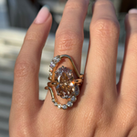 A close-up of a hand wearing a large rose gold ring with a prominent oval champagne-colored center stone and two white diamond wave stacking bands