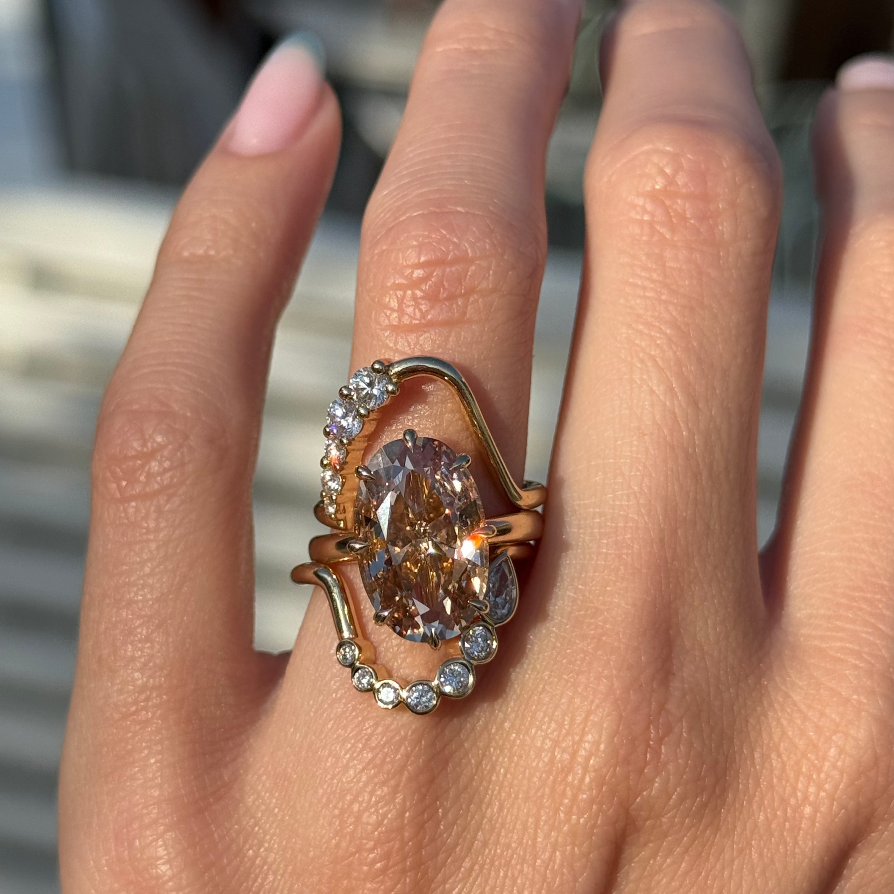 A close-up of a hand wearing a large rose gold ring with a prominent oval champagne-colored center stone and two white diamond wave stacking bands