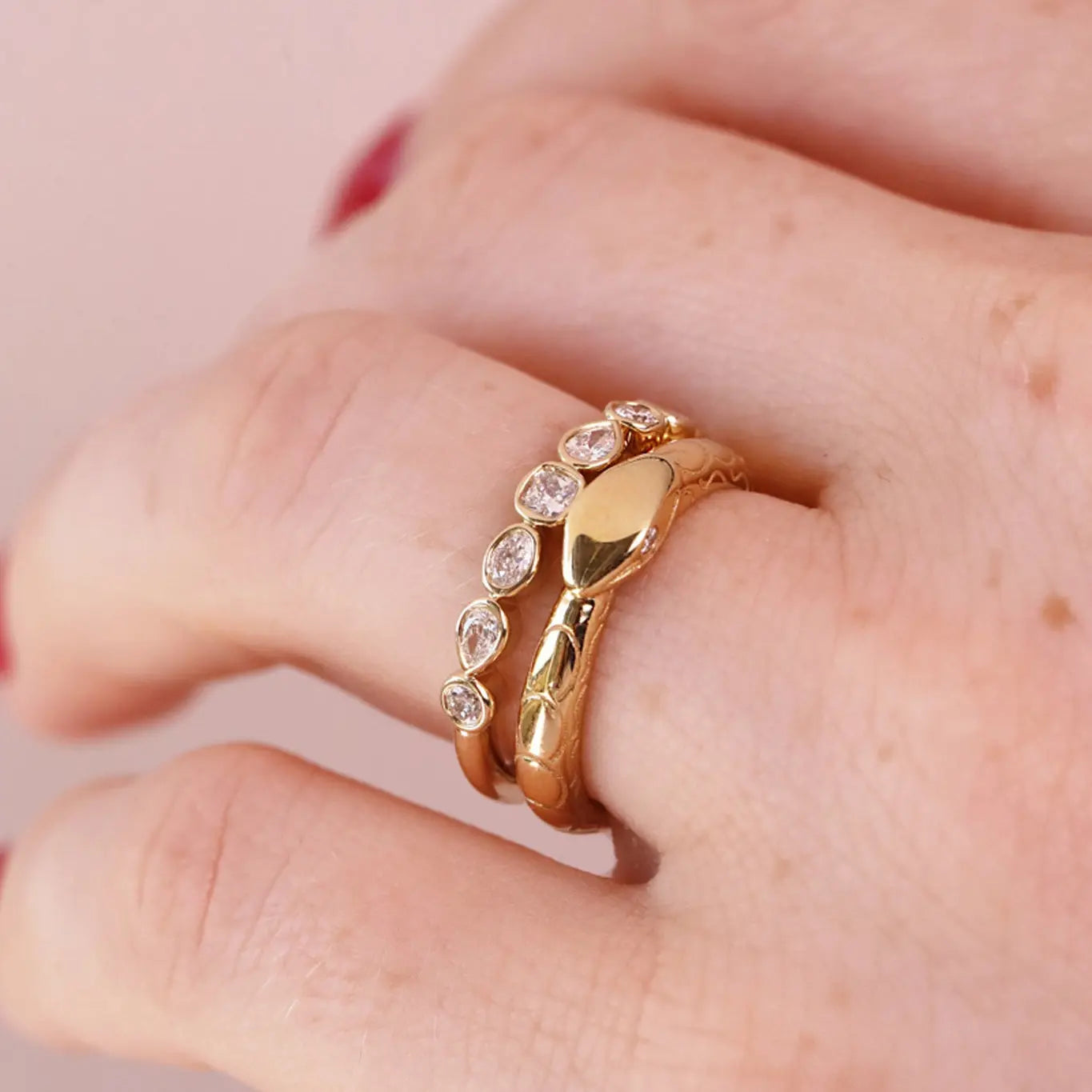 A close-up of a hand wearing a gold snake ring with small clear stones along its body, set against a soft pink background.
