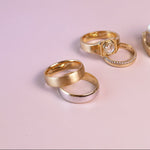 Four rings displayed on a soft pink background, including two gold rings, one silver ring, and a pair stacked on a small gold block. One ring features a clear central stone.