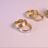 Four rings displayed on a soft pink background, including two gold rings, one silver ring, and a pair stacked on a small gold block. One ring features a clear central stone.
