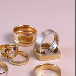 A set of six rings in various styles and metals, including yellow gold, silver, and a two-tone gold and silver band, arranged on a soft pink background.