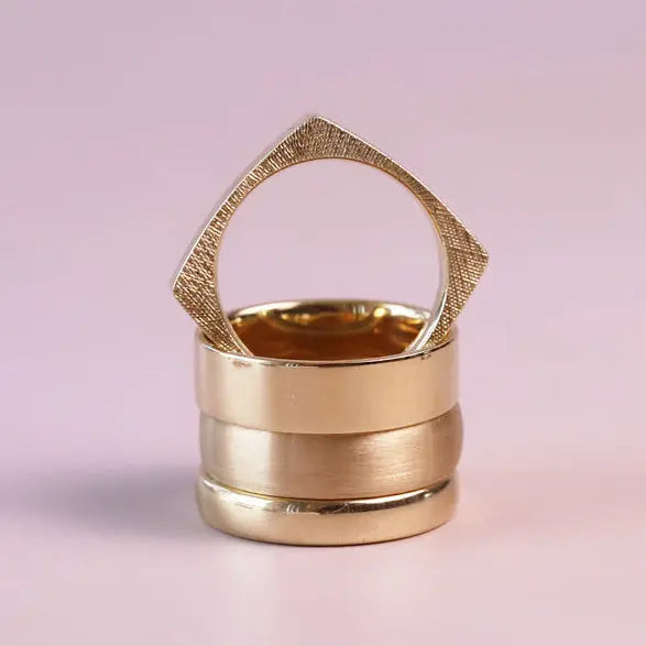 A gold-toned ring with a textured triangular top and stacked cylindrical bands, photographed on a soft light pink background.