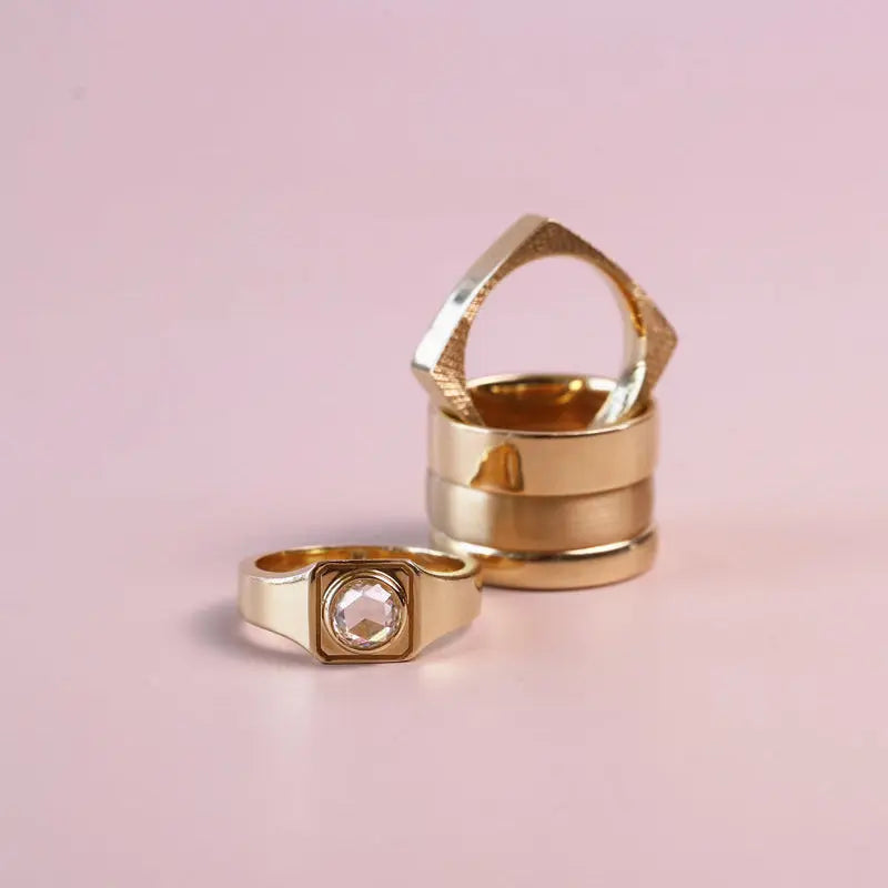 Two gold rings on a soft pink background, featuring a stacked geometric ring and a square bezel-set ring with a clear crystal.