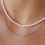 A close-up of a person's neck and upper chest wearing two layered necklaces: one white pearl strand and one delicate gold paperclip chain.