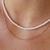A close-up of a person's neck and upper chest wearing two layered necklaces: one white pearl strand and one delicate gold paperclip chain.