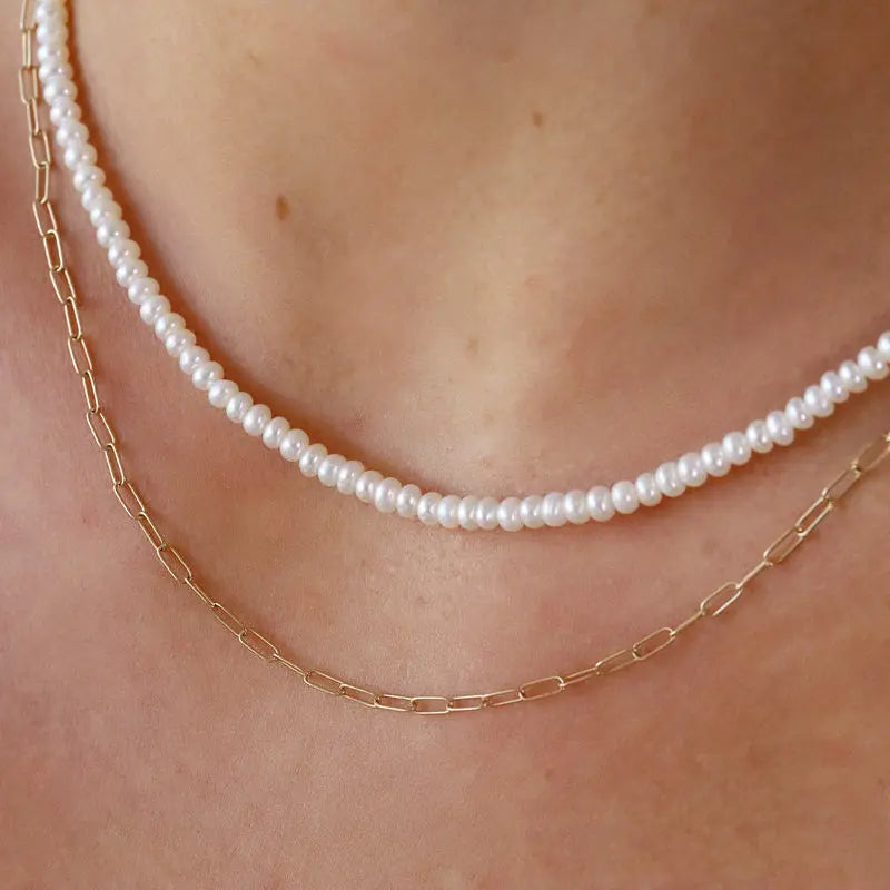 A close-up of a person's neck and upper chest wearing two layered necklaces: one white pearl strand and one delicate gold paperclip chain.