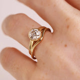 A close-up of a hand wearing a gold ring with a round-cut clear gemstone in a bezel setting, photographed against a soft light pink background.