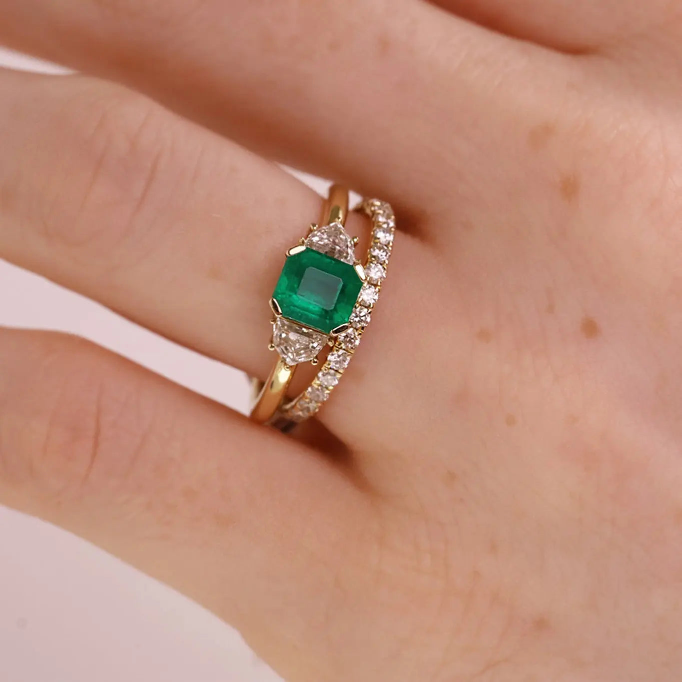 A close-up of a hand wearing a gold ring with a square-cut green emerald gemstone and pavé-set clear stones along the band.