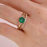 A close-up of a hand wearing a gold ring with a square-cut green emerald gemstone and pavé-set clear stones along the band.