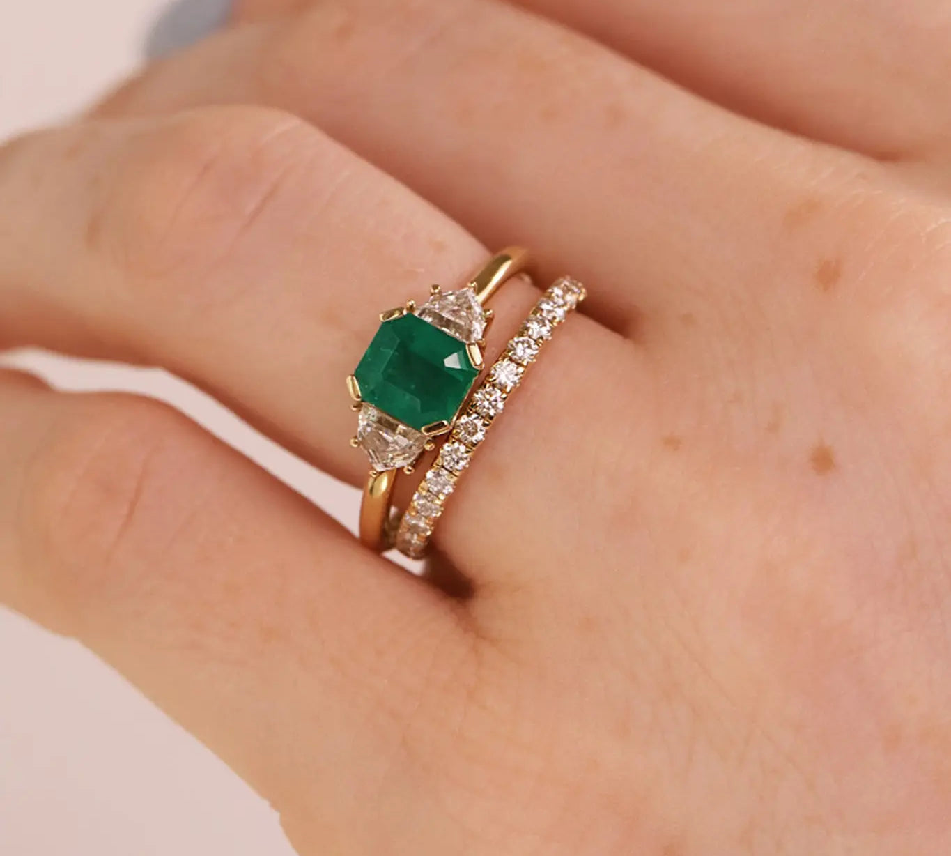 A close-up of a hand wearing a gold ring with a square-cut green emerald center stone and a second slim gold band set with small clear stones.