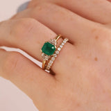 A close-up of a hand wearing a gold ring with a square-cut green emerald center stone and a second slim gold band set with small clear stones.