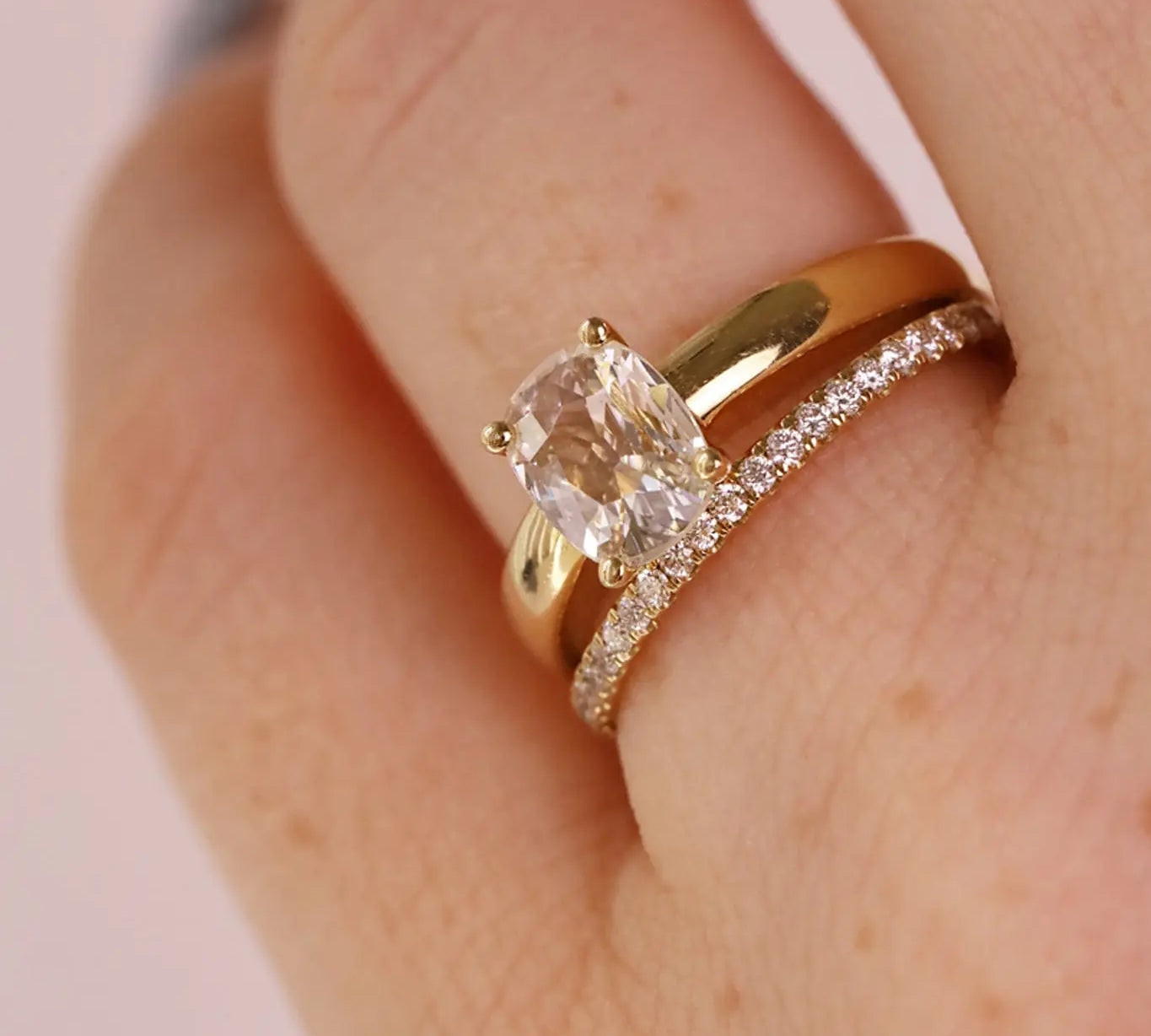 A close-up of a hand wearing a gold engagement ring with a cushion-cut clear gemstone and a matching gold wedding band with small pavé stones, against a soft pink background.