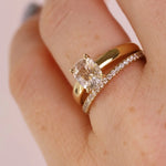 A close-up of a hand wearing a gold engagement ring with a cushion-cut clear gemstone and a matching gold wedding band with small pavé stones, against a soft pink background.