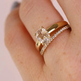 A close-up of a hand wearing a gold engagement ring with a cushion-cut clear gemstone and a matching gold wedding band with small pavé stones, against a soft pink background.