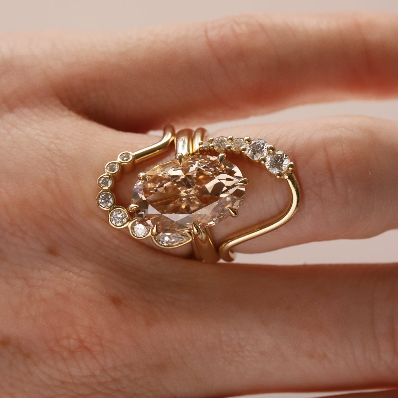 A gold ring with a large peach-colored heart-shaped center stone and small clear accent stones, shown worn on a hand against a soft beige background.