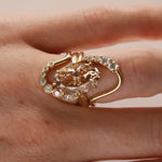 A gold ring with a large peach-colored heart-shaped center stone and small clear accent stones, shown worn on a hand against a soft beige background.