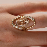 A gold ring with a large peach-colored heart-shaped center stone and small clear accent stones, shown worn on a hand against a soft beige background.