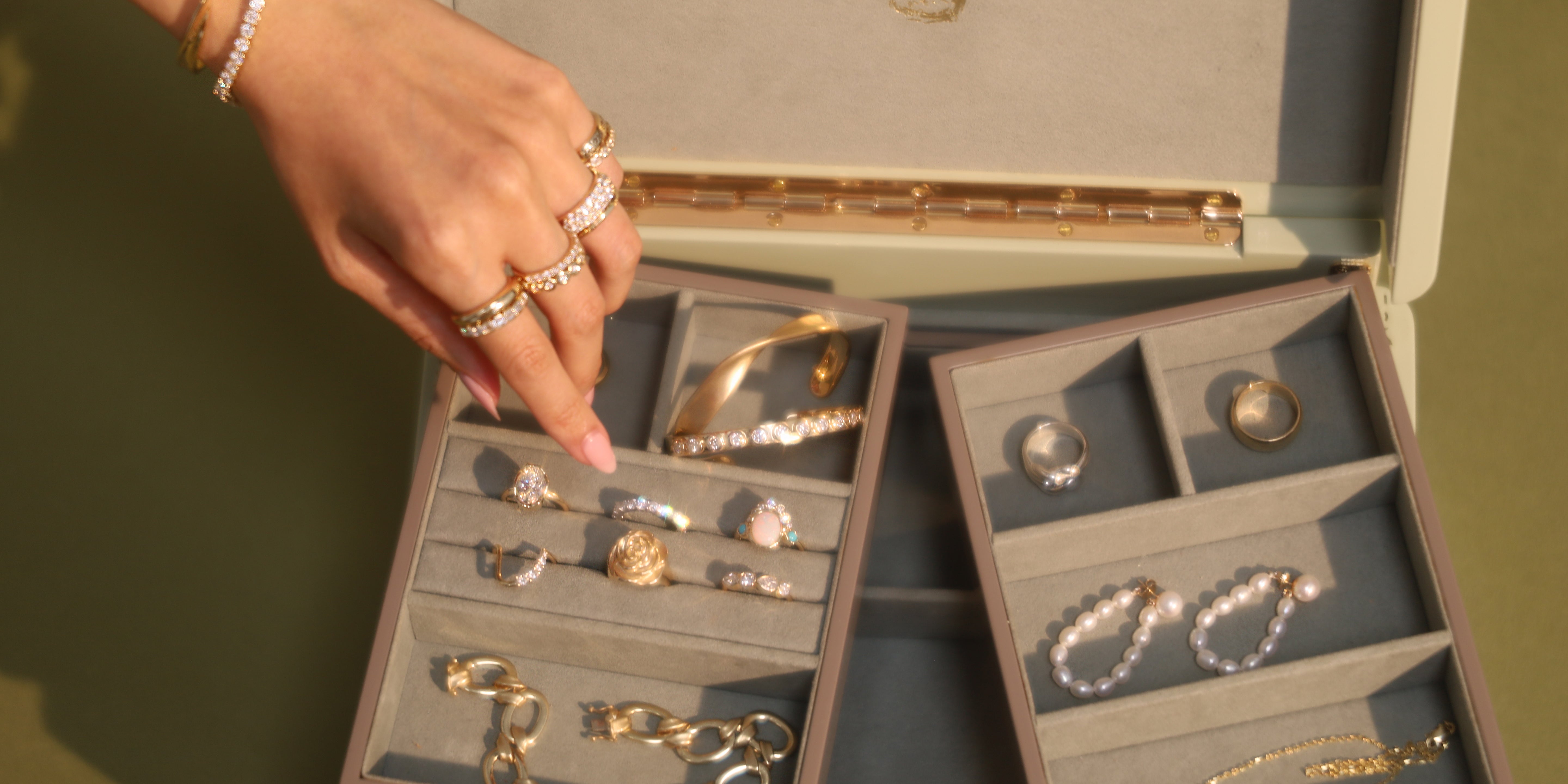 Hand reaching into open green jewelry box with gold, pearl and diamond jewelry inside on green background