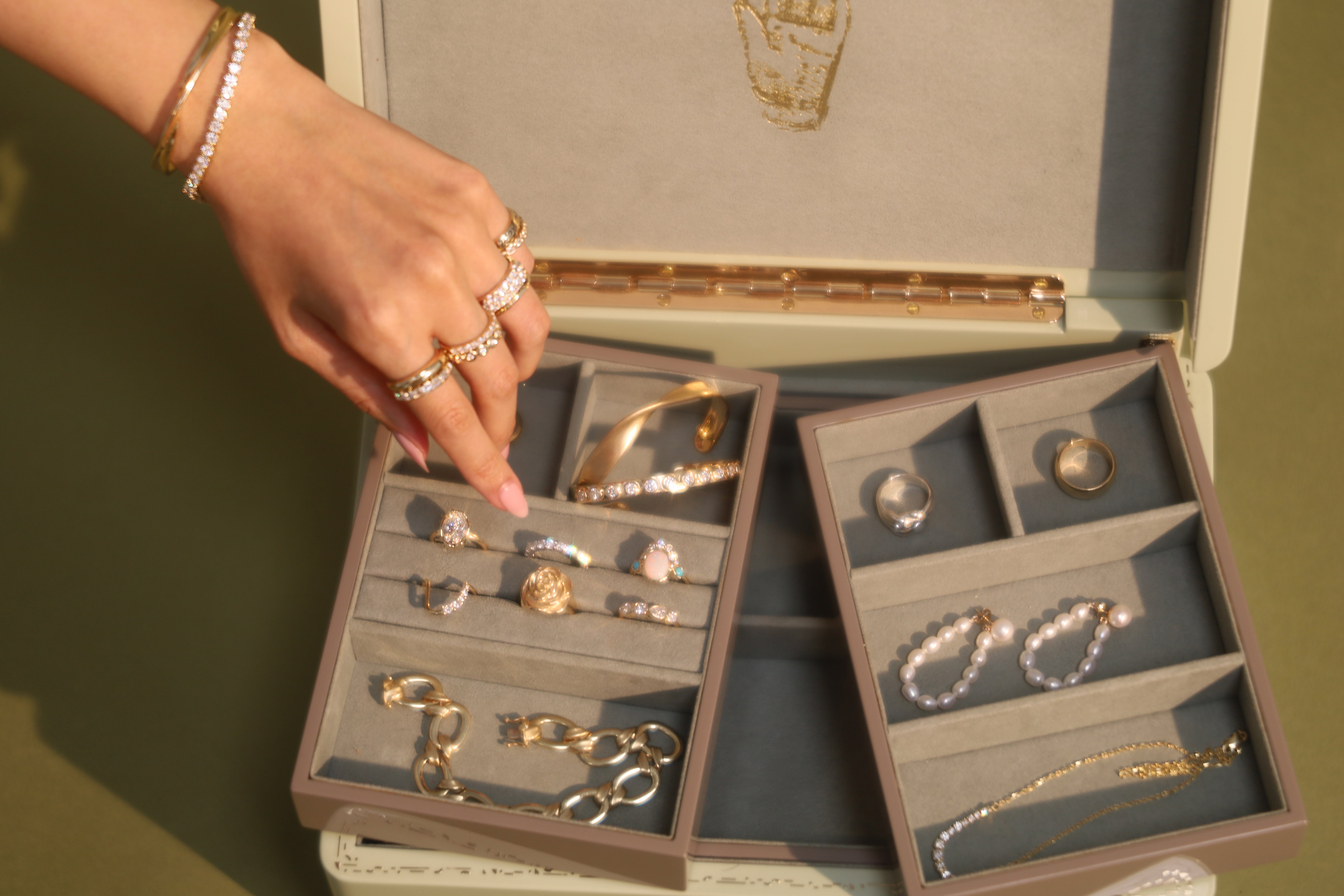 Hand reaching into open green jewelry box with gold, pearl and diamond jewelry inside on green background