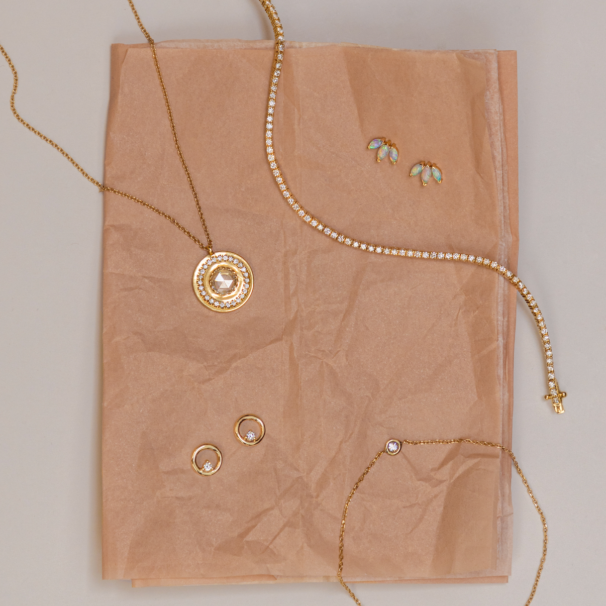 Flat lay of yellow gold fine jewelry on crumpled blush tissue paper: White Diamond La Femme Amulet, Coco Tennis Bracelet, Opal Lotus Studs, White Diamond Orbit Studs, Dainty Diamond Bezel Bracelet.