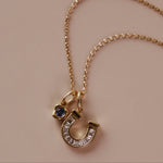 A gold chain necklace with a horseshoe-shaped charm stacked with a bezel gemstone charm, displayed on a solid beige background.