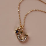 A gold chain necklace with a horseshoe-shaped charm stacked with a bezel gemstone charm, displayed on a solid beige background.