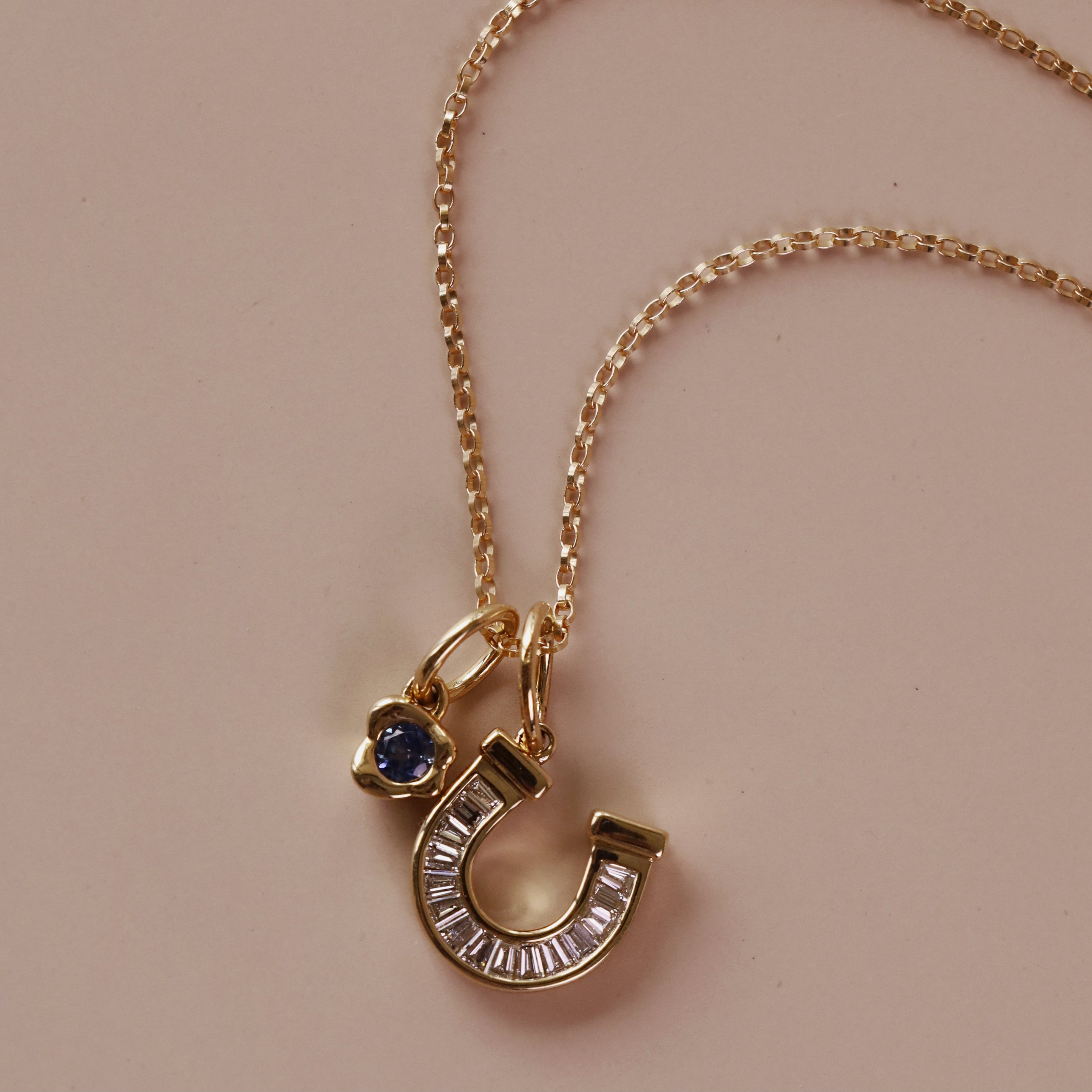 A gold chain necklace with a horseshoe-shaped charm stacked with a bezel gemstone charm, displayed on a solid beige background.