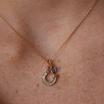 A close-up of a person's neck wearing a delicate gold chain necklace with a horseshoe-shaped pendant stacked with a  gemstone bezel charm.