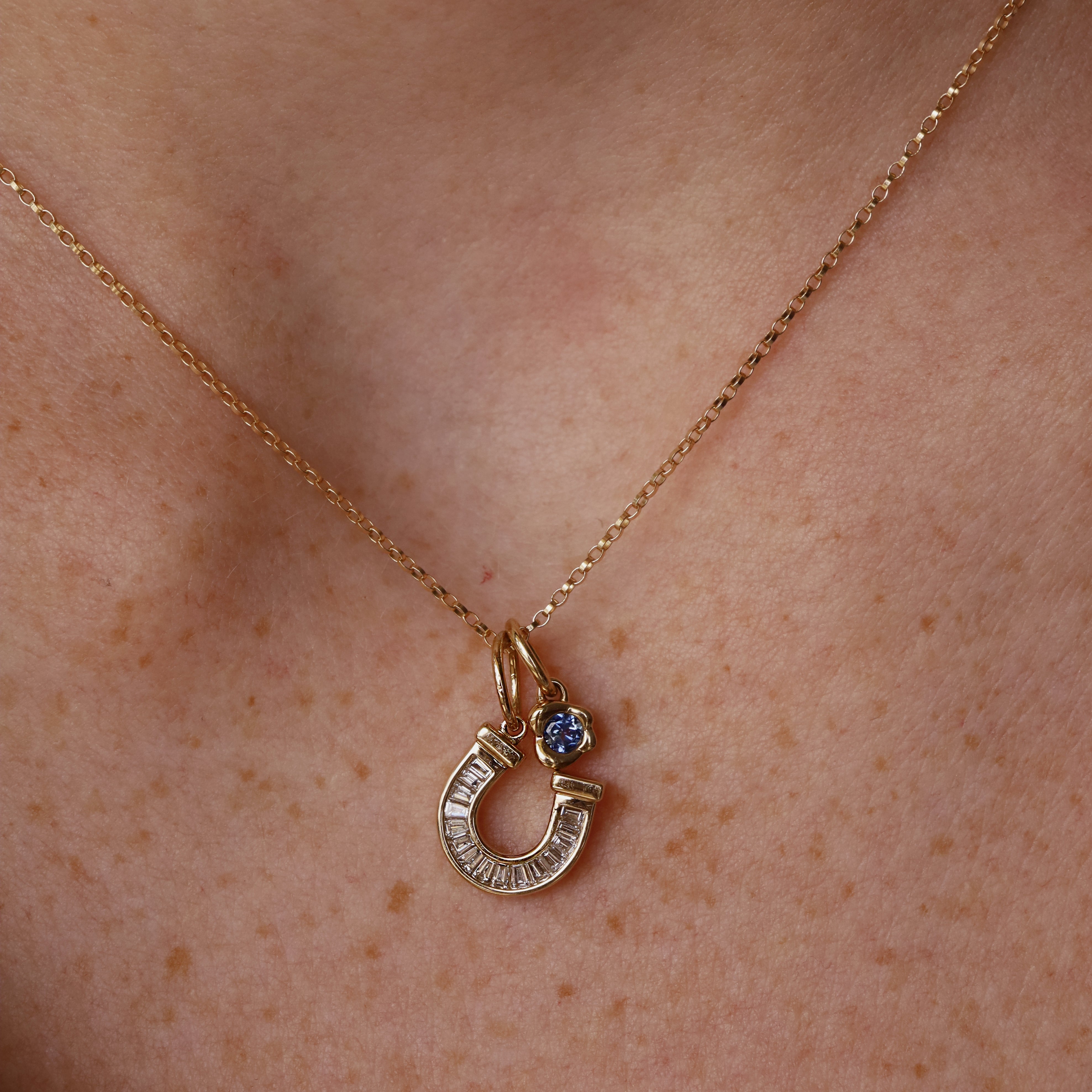 A close-up of a person's neck wearing a delicate gold chain necklace with a horseshoe-shaped pendant stacked with a  gemstone bezel charm.