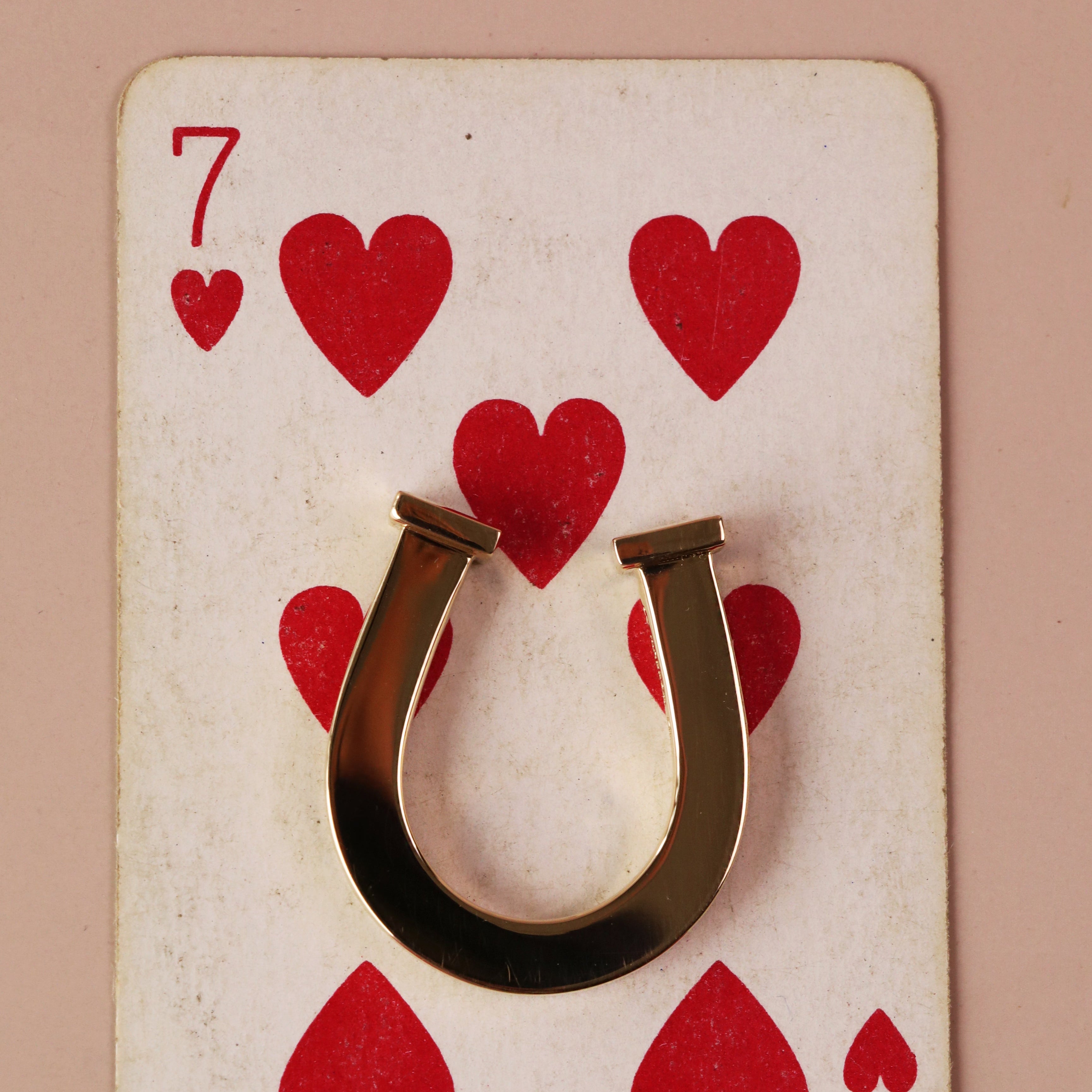 A gold horseshoe-shaped ring or pendant displayed on a vintage-style 7 of hearts playing card against a solid dusty pink background.
