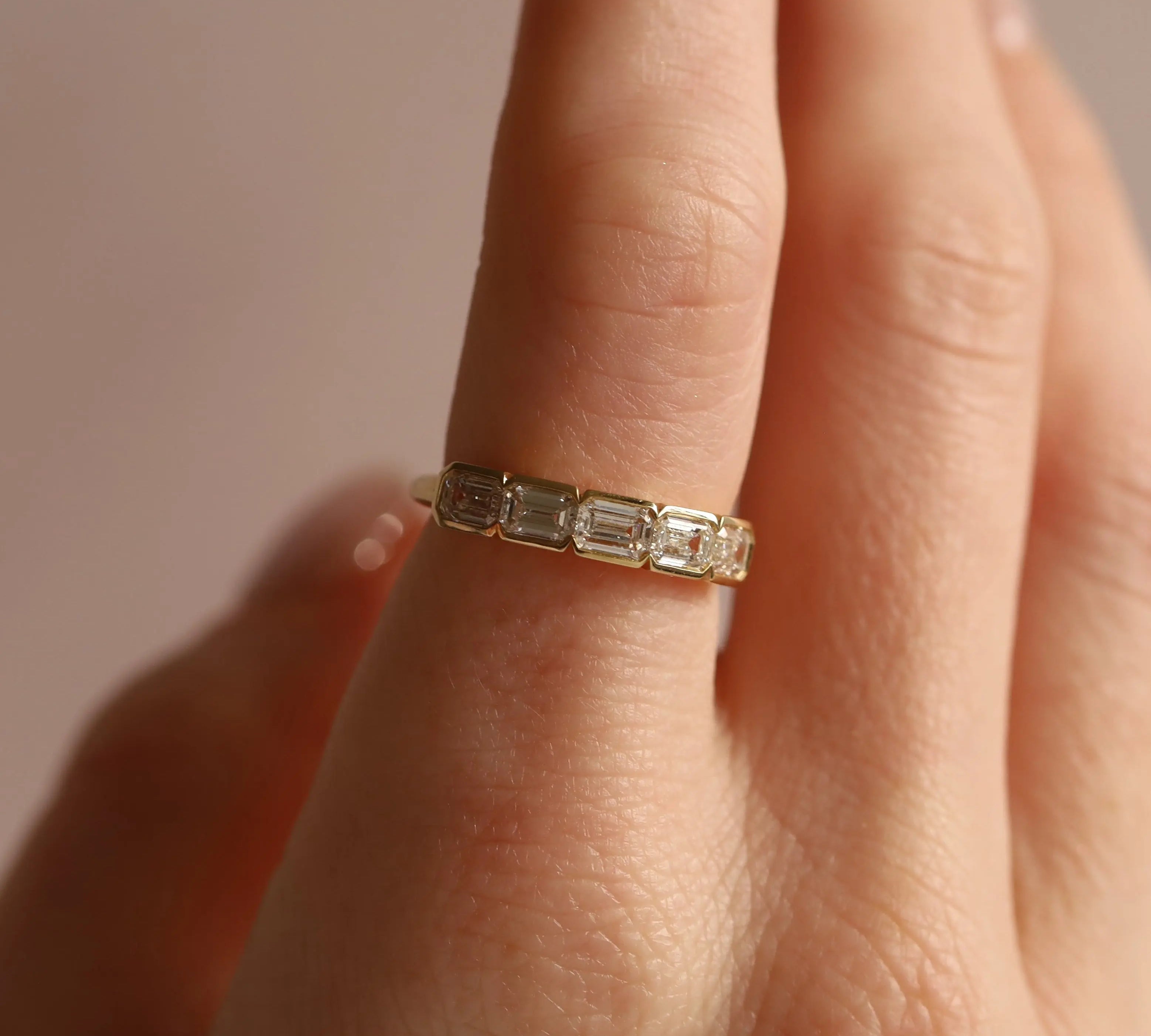 Gold ring with emerald-cut diamonds on a finger against a neutral background