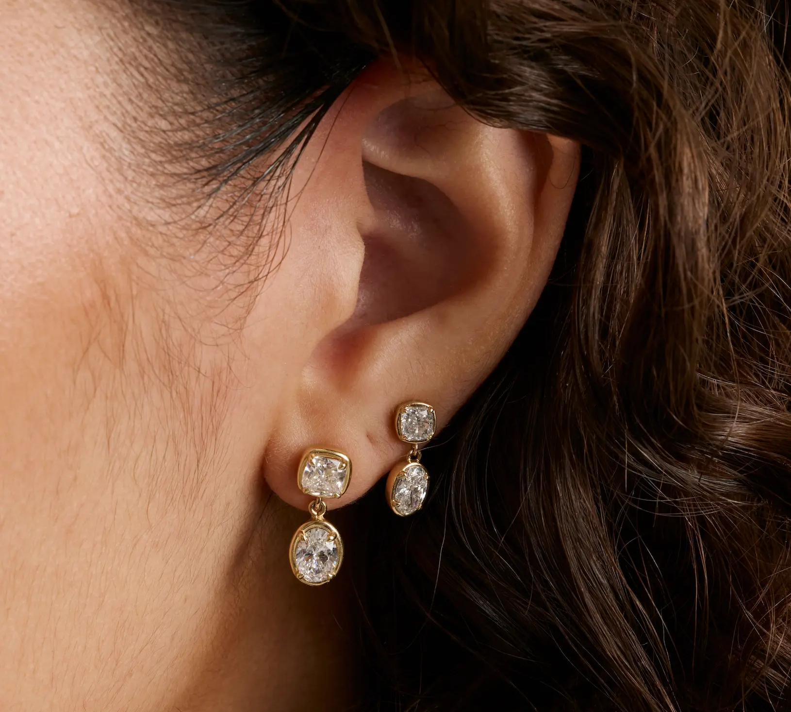 Close up of woman's ear with curly brown hair showing two diamond drop earrings - Large Lisse Dangle Earrings and Lisse Dangle Earrings
