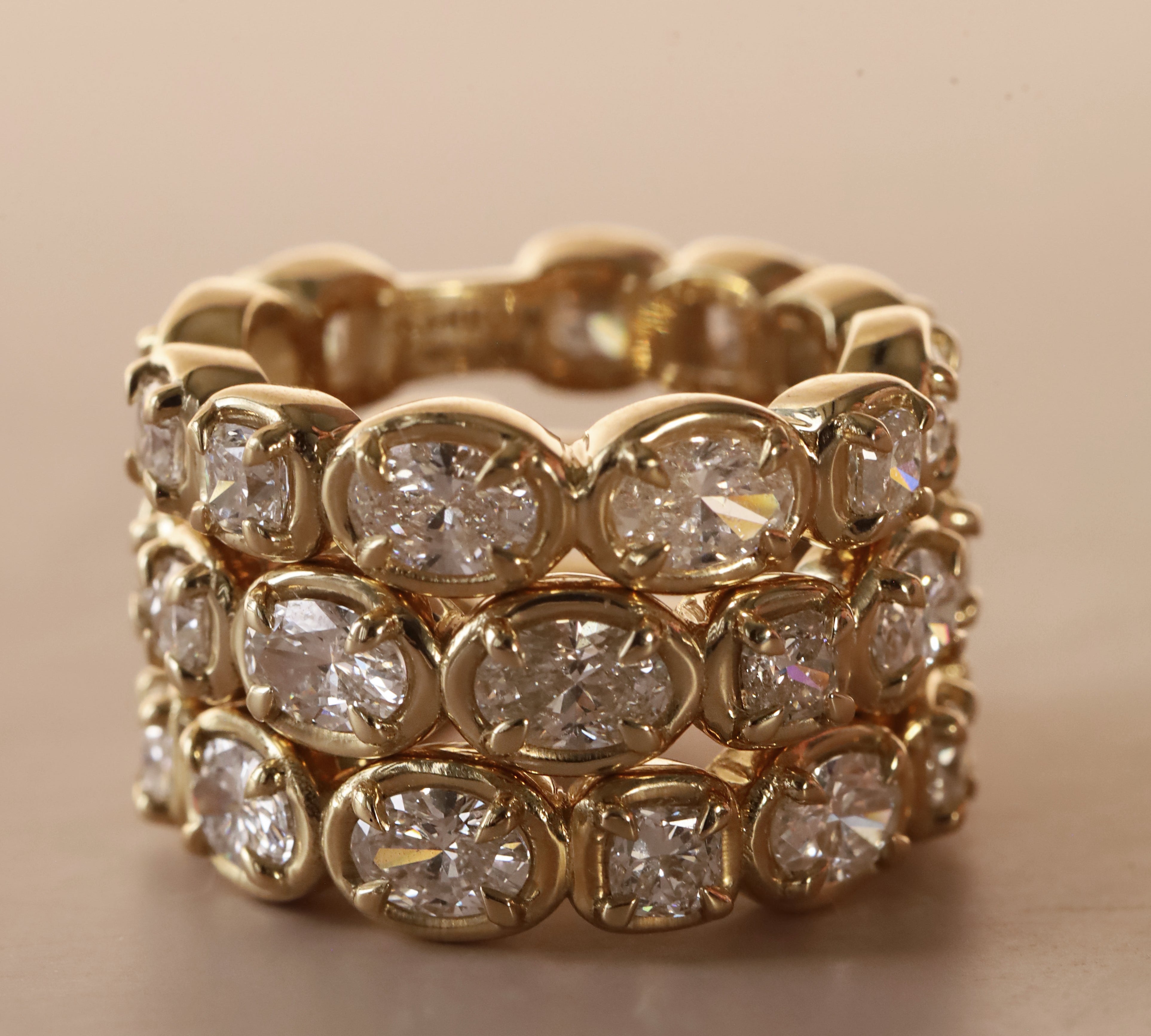 Straight diamond band featuring alternating oval and cushion cut diamonds, set in yellow gold. 3 of them stacked together against a neutral background.