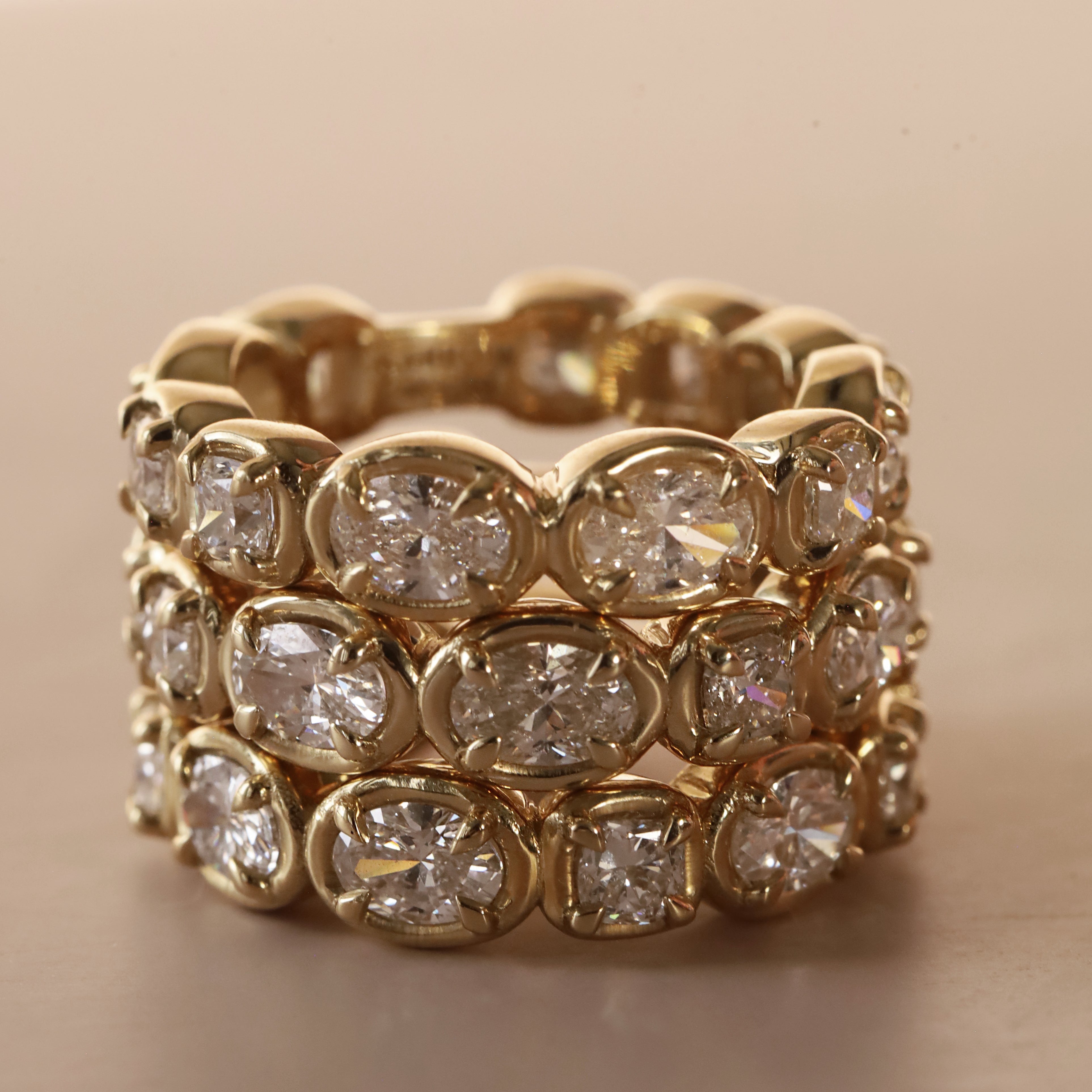 Straight diamond band featuring alternating oval and cushion cut diamonds, set in yellow gold. 3 of them stacked together against a neutral background.