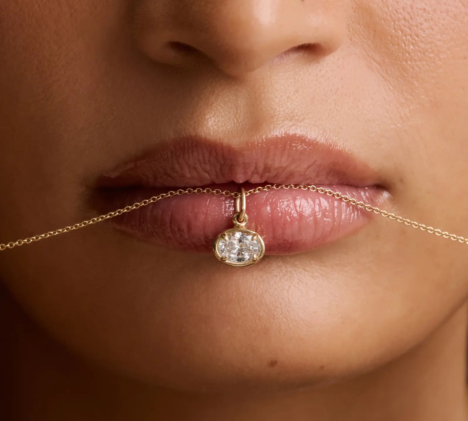 Close-up of a model's lips holding a delicate gold chain necklace with a small oval diamond pendant.