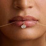 Close-up of a model's lips holding a delicate gold chain necklace with a small oval diamond pendant.