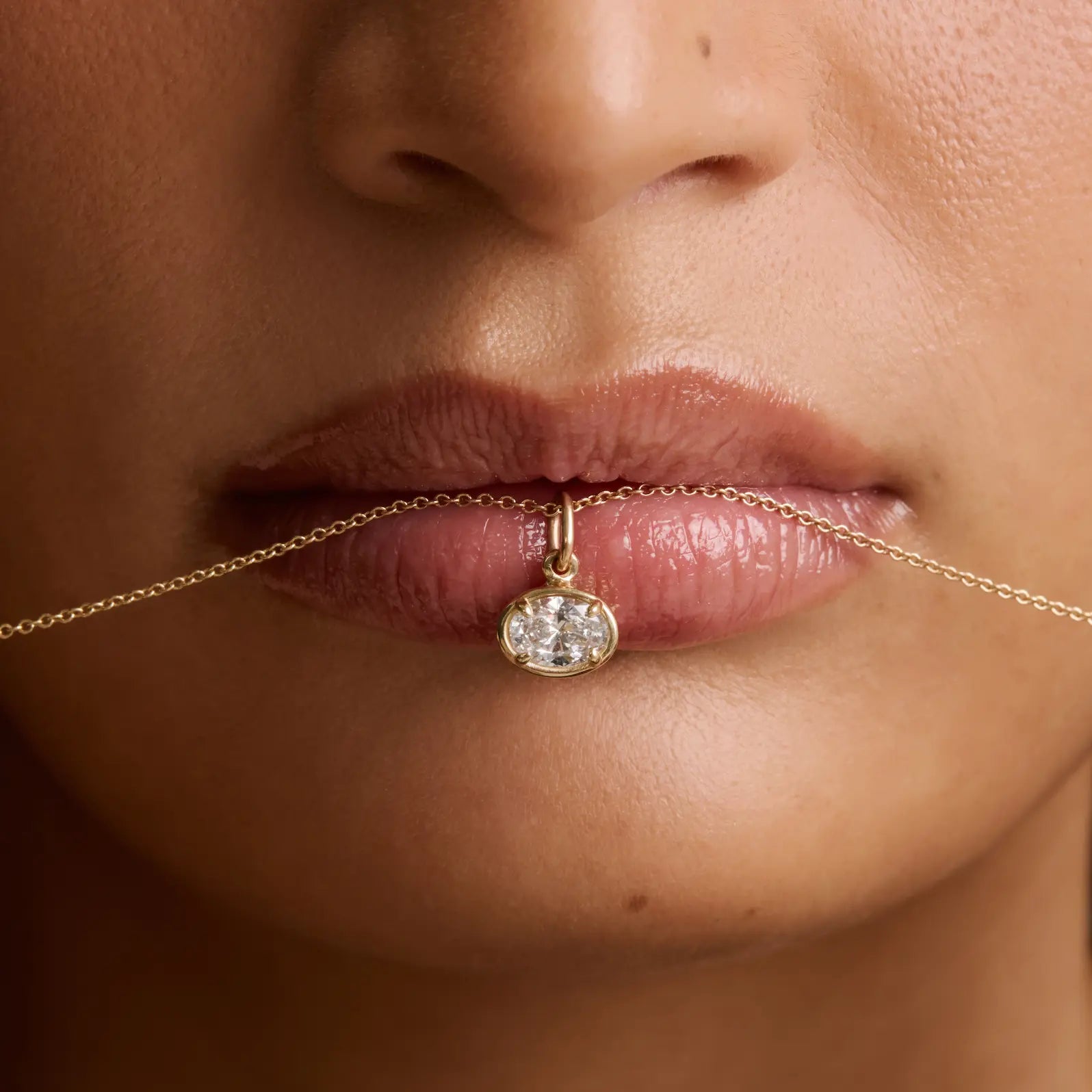 Close-up of a model's lips holding a delicate gold chain necklace with a small oval diamond pendant.