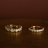 Two gold eternity rings set with round-cut clear gemstones, displayed side-by-side on a dark brown reflective surface against a matching dark brown background.